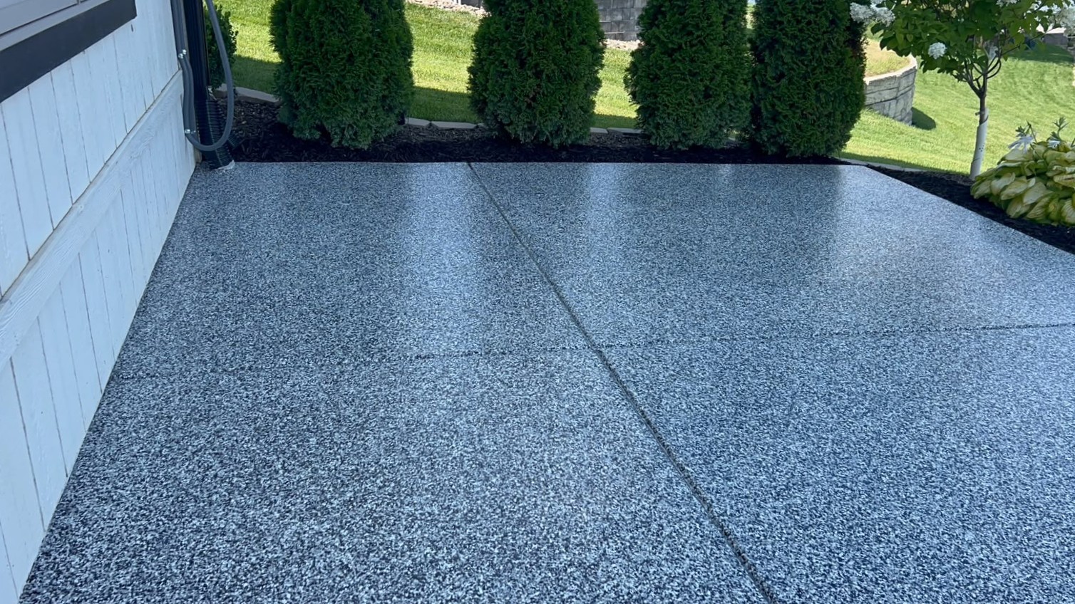 Patio floor coating with expansion joints, white siding, and evergreen trees at the far edge